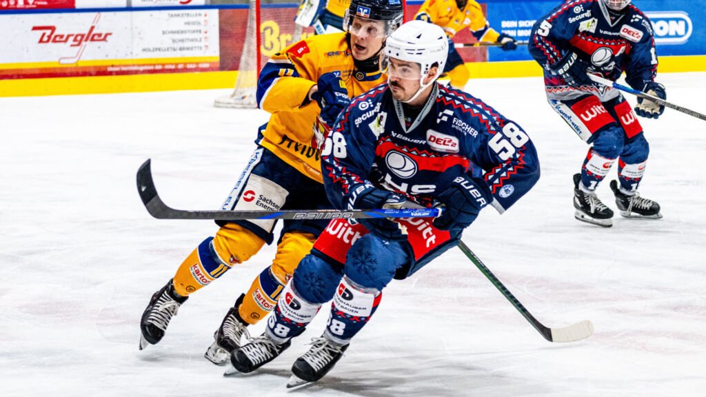 Zwei Eishockeyspieler, Charlie Jahnke (links) und Alex-Olivier Voyer (rechts) kämpfen auf dem Eis um die Scheibe: Voyer im blauen Trikot mit der Nummer 58 fährt nach vorne, während Jahnke im gelben Trikot dicht hinter ihm versucht, ihn zu stoppen. Im Hintergrund sind weitere Spieler und die Bande der Hans-Schröpf-Arena in Weiden zu sehen.