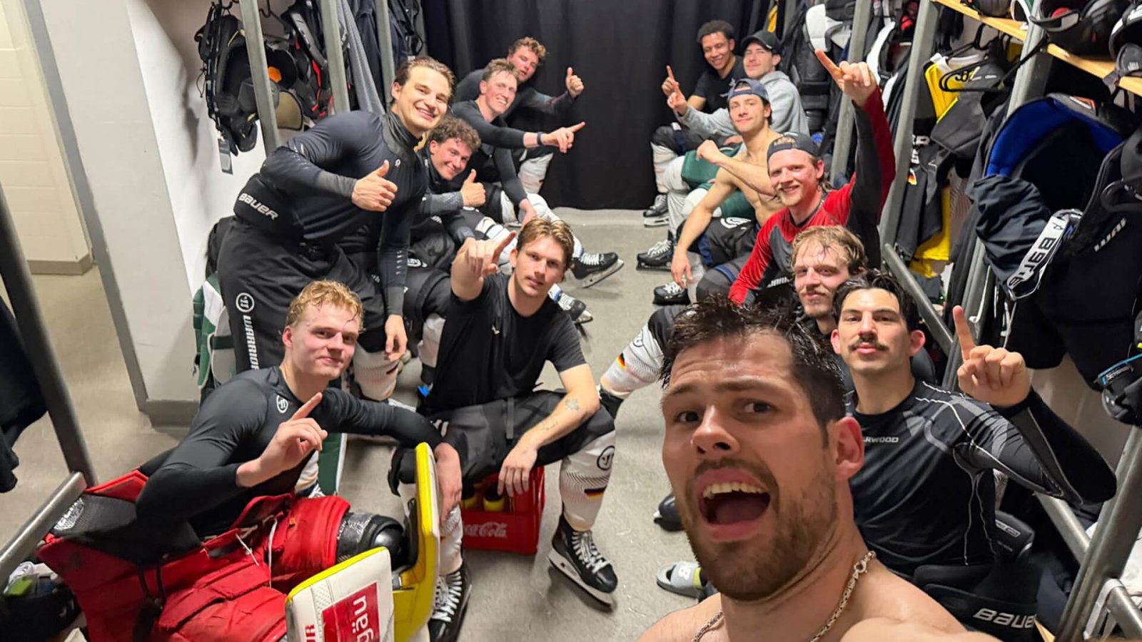 Eishockey-Team feiert gemeinsam in der Kabine und posiert nach dem Spiel mit erhobenen Fingern und Daumen in die Kamera.