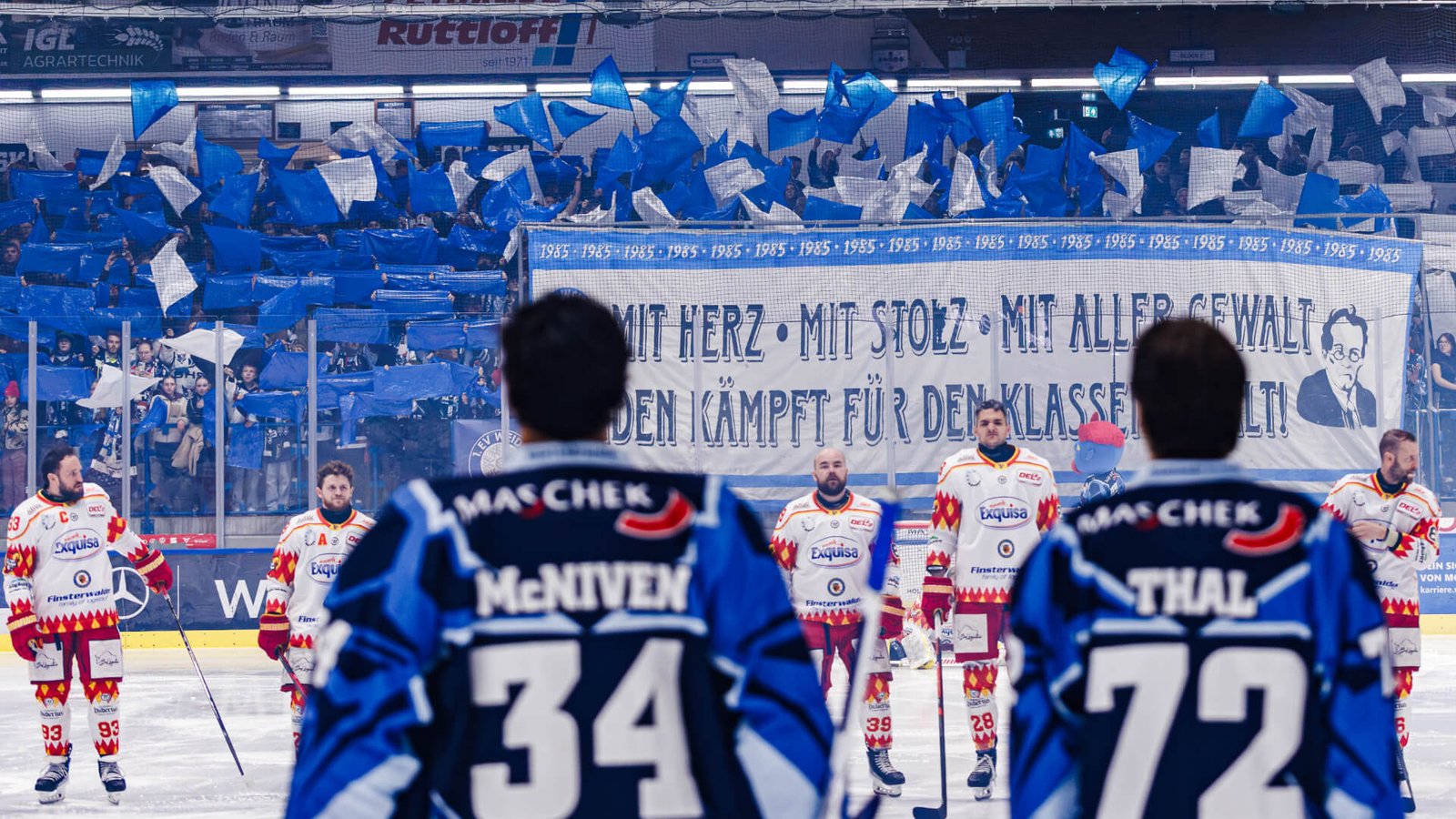 Eishockeyspieler stehen auf dem Eis vor einer Fankurve, in der Fans blaue und weiße Fahnen schwenken und ein großes Banner präsentieren.