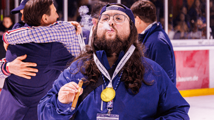 Christian mit Medaille auf dem Eis der Blue Devils Weiden, mit Zigarre in der Hand und feiernder Atmosphäre im Hintergrund.
