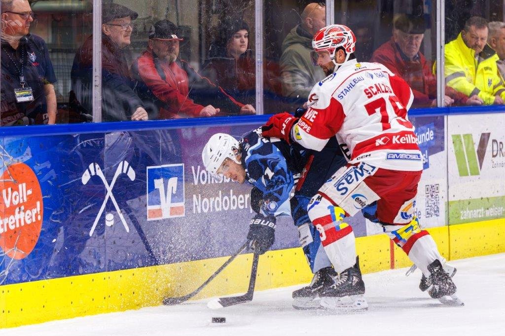 Eishockeyspieler in rotem Trikot checkt einen Gegenspieler in blauem Trikot an der Bande, während Zuschauer hinter der Plexiglasscheibe zusehen.