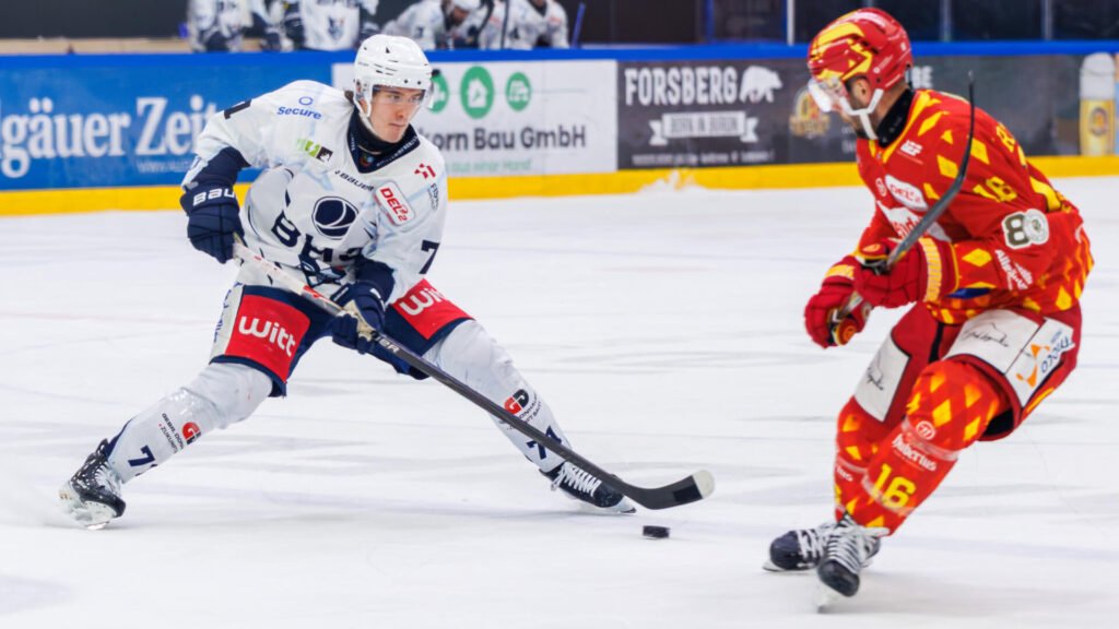 Zwei Eishockeyspieler kämpfen auf dem Eis um den Puck: Cedric Schiemenz im weißen Trikot führt den Puck mit dem Schläger, während ein Gegenspieler im rot-gelben Trikot auf ihn zufährt.