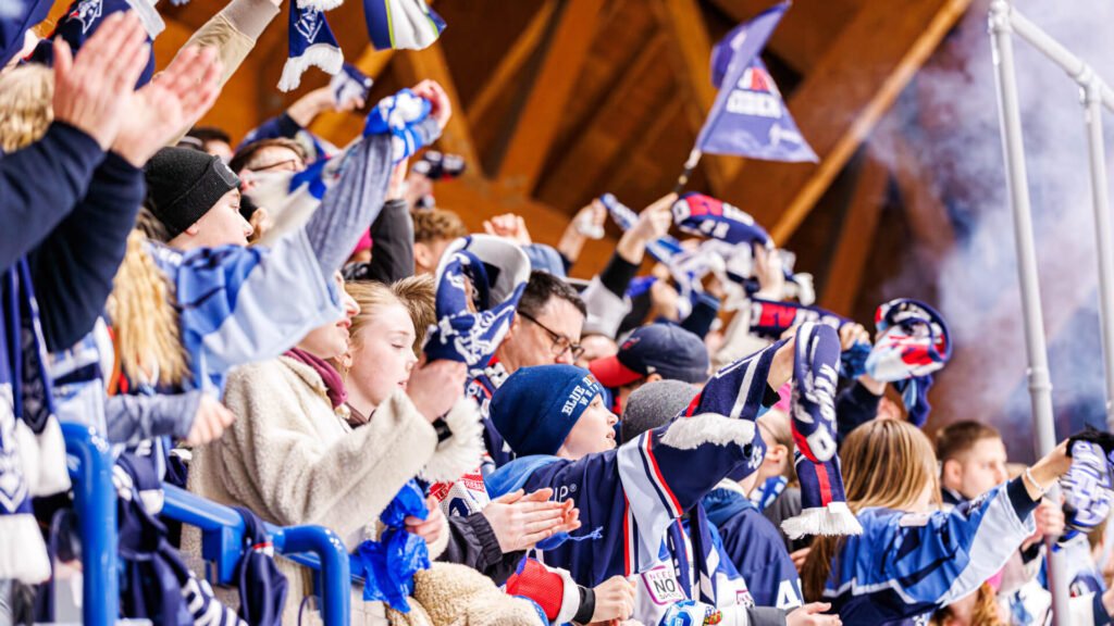 Eishockey-Fans feiern auf der Tribüne und schwenken blau-weiße Fan-Schals.